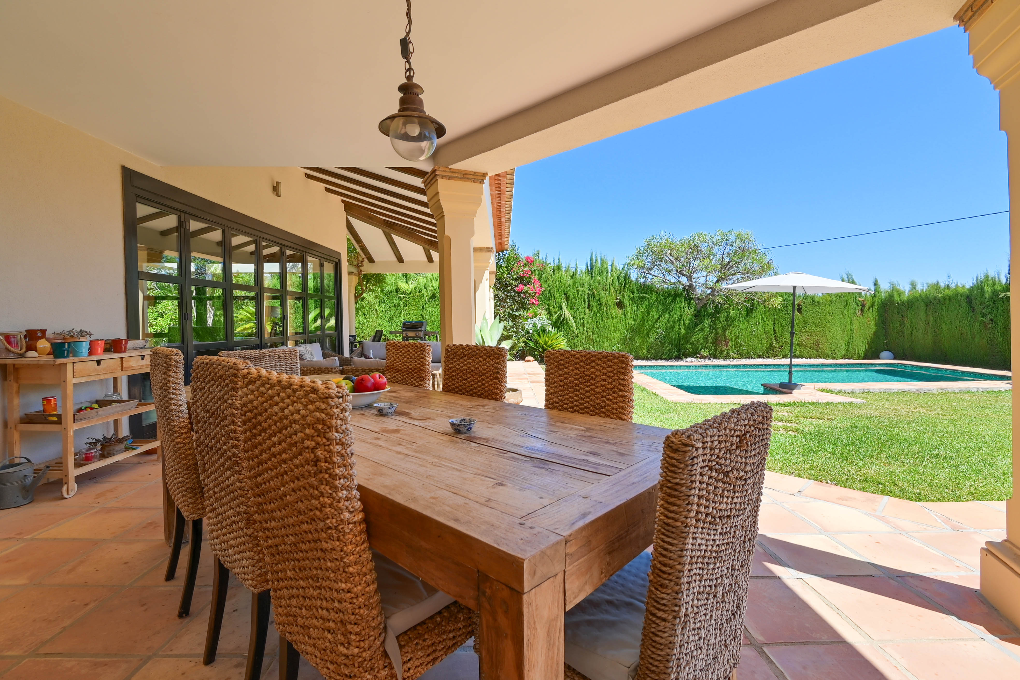 Salle à manger extérieure couverte avec table en bois massif et chaises en rotin donnant sur la piscine et le jardin.
