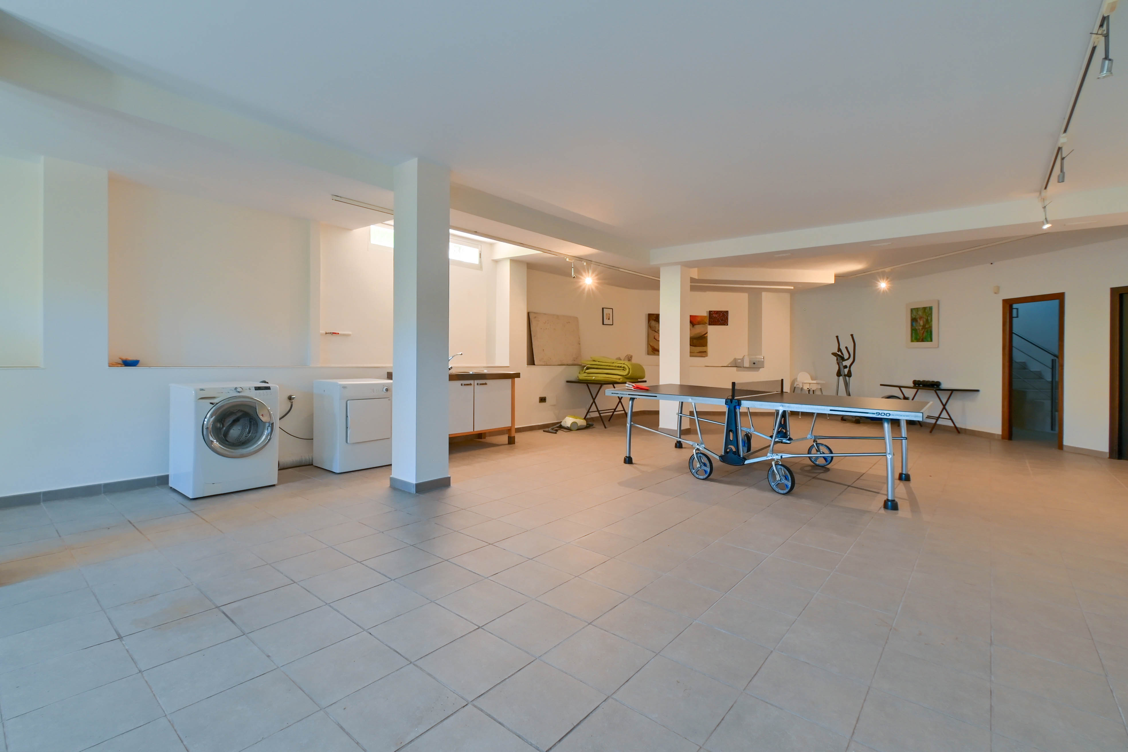 Salle de jeux spacieuse avec lave-linge, sèche-linge et table de ping-pong sur carrelage couleur crème.