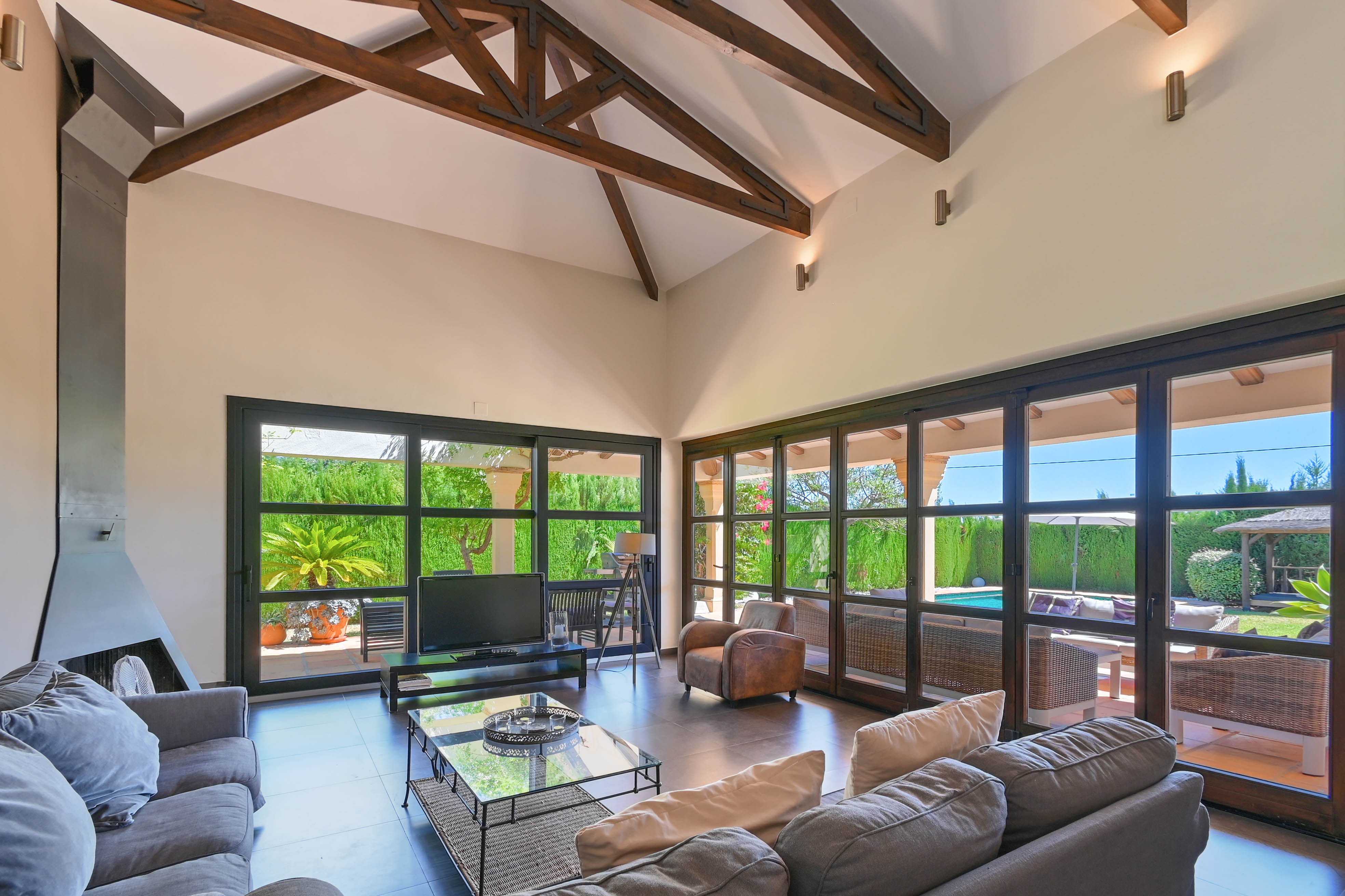 Salon lumineux avec poutres apparentes, cheminée moderne et vue sur le jardin avec piscine à travers de larges fenêtres.