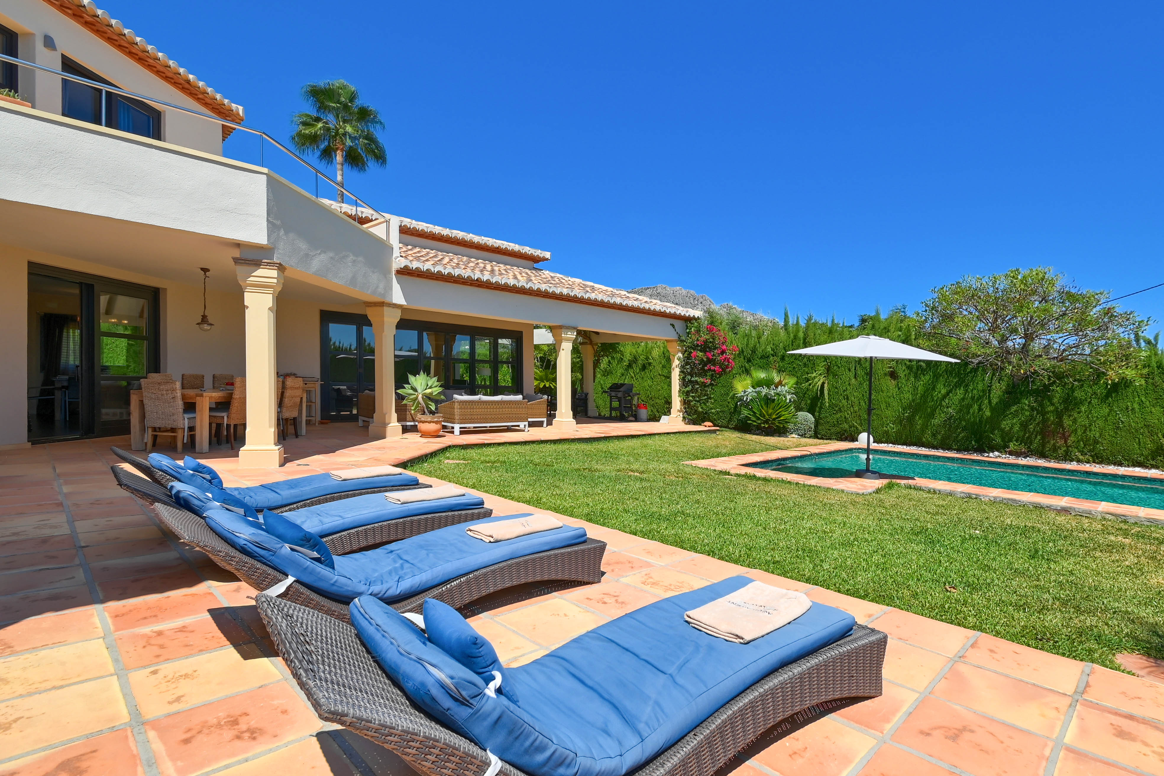 Terrasse avec chaises longues bleues et piscine allongée dans un jardin gazonné.