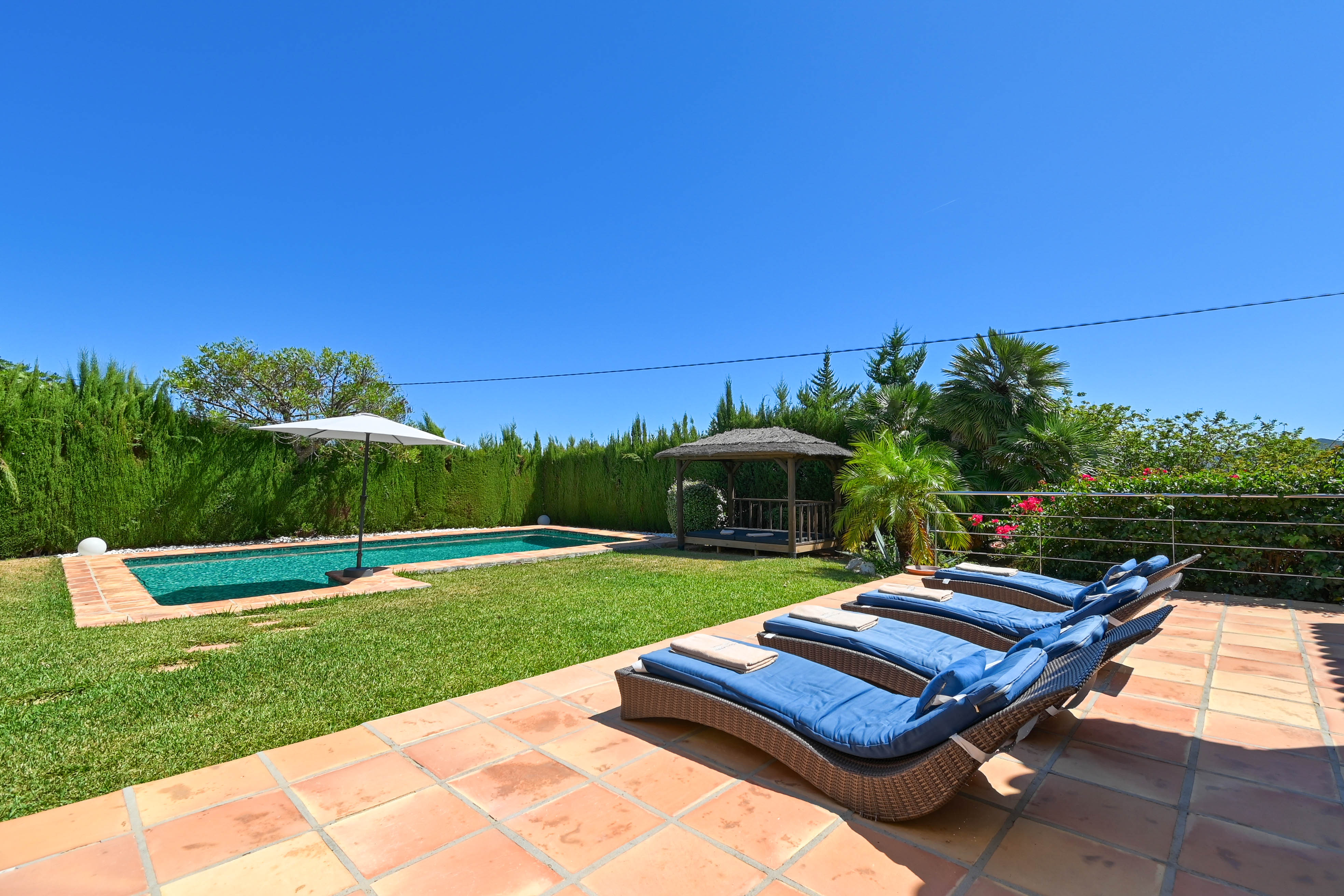 Terrasse ensoleillée avec transats en rotin bleu, piscine et gazebo dans un jardin paysager.