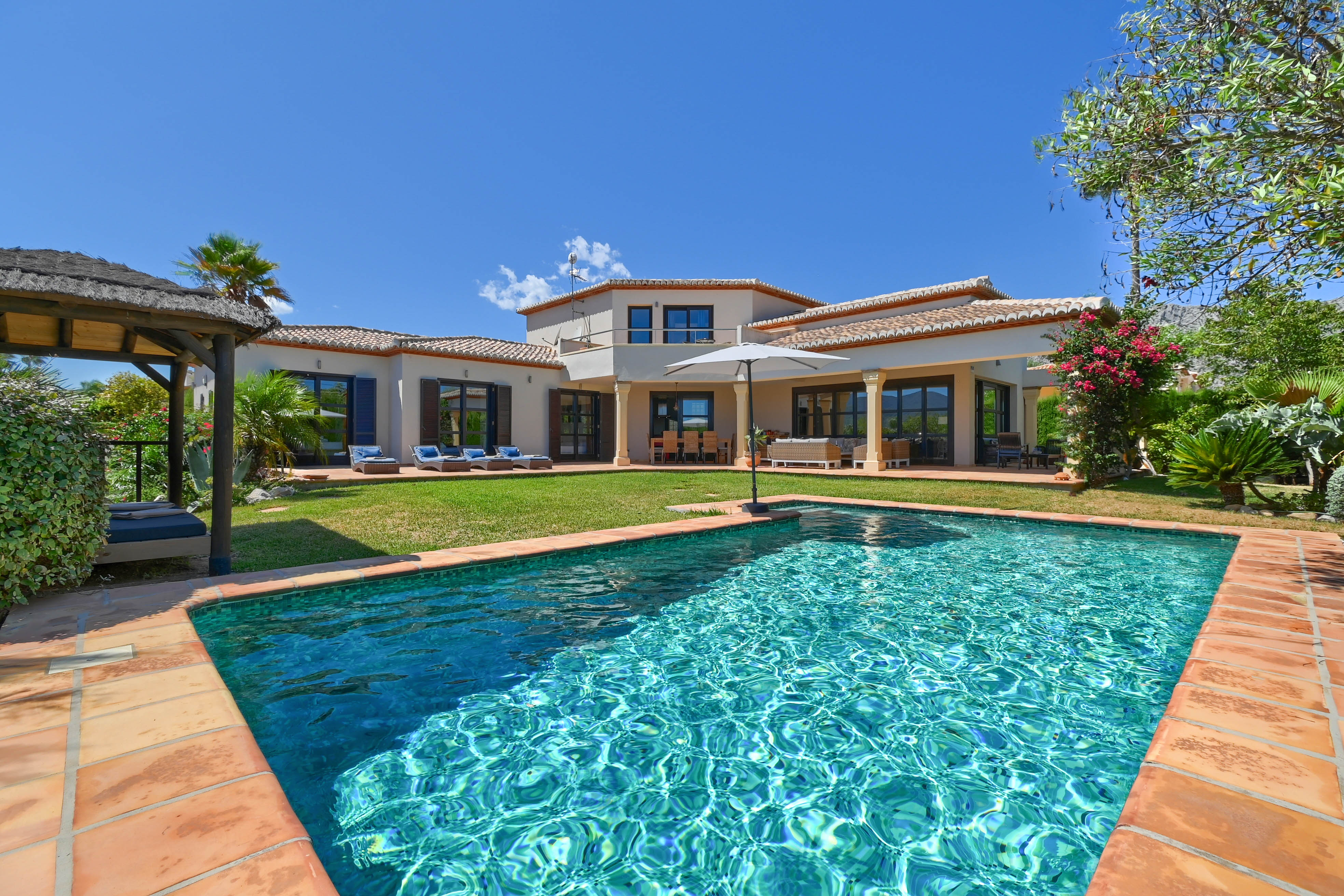 Vue de la piscine turquoise bordée de terre cuite et de la villa à deux étages avec volets.