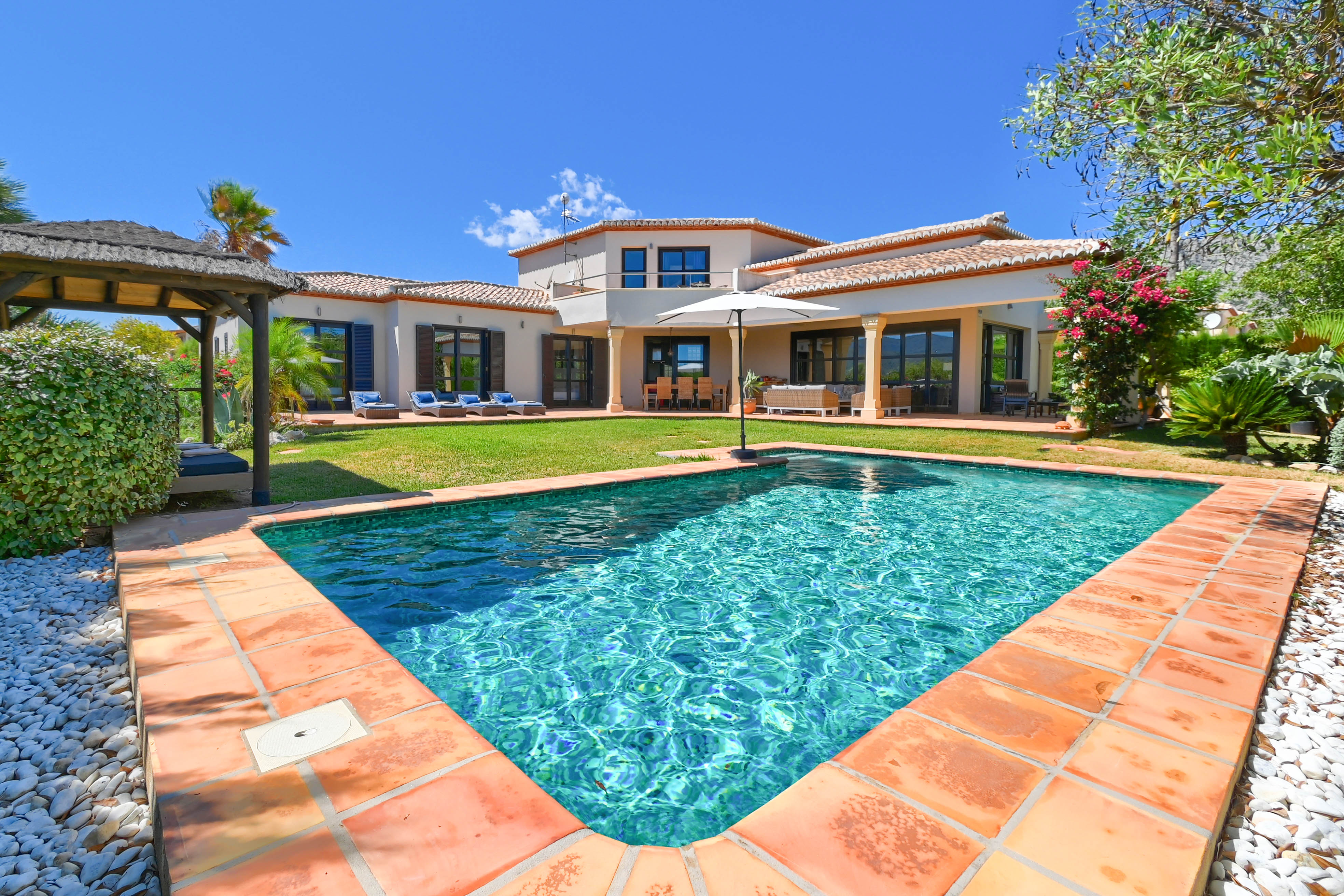 Vue sur la piscine turquoise bordée de terre cuite et la villa avec jardin et gazebo.
