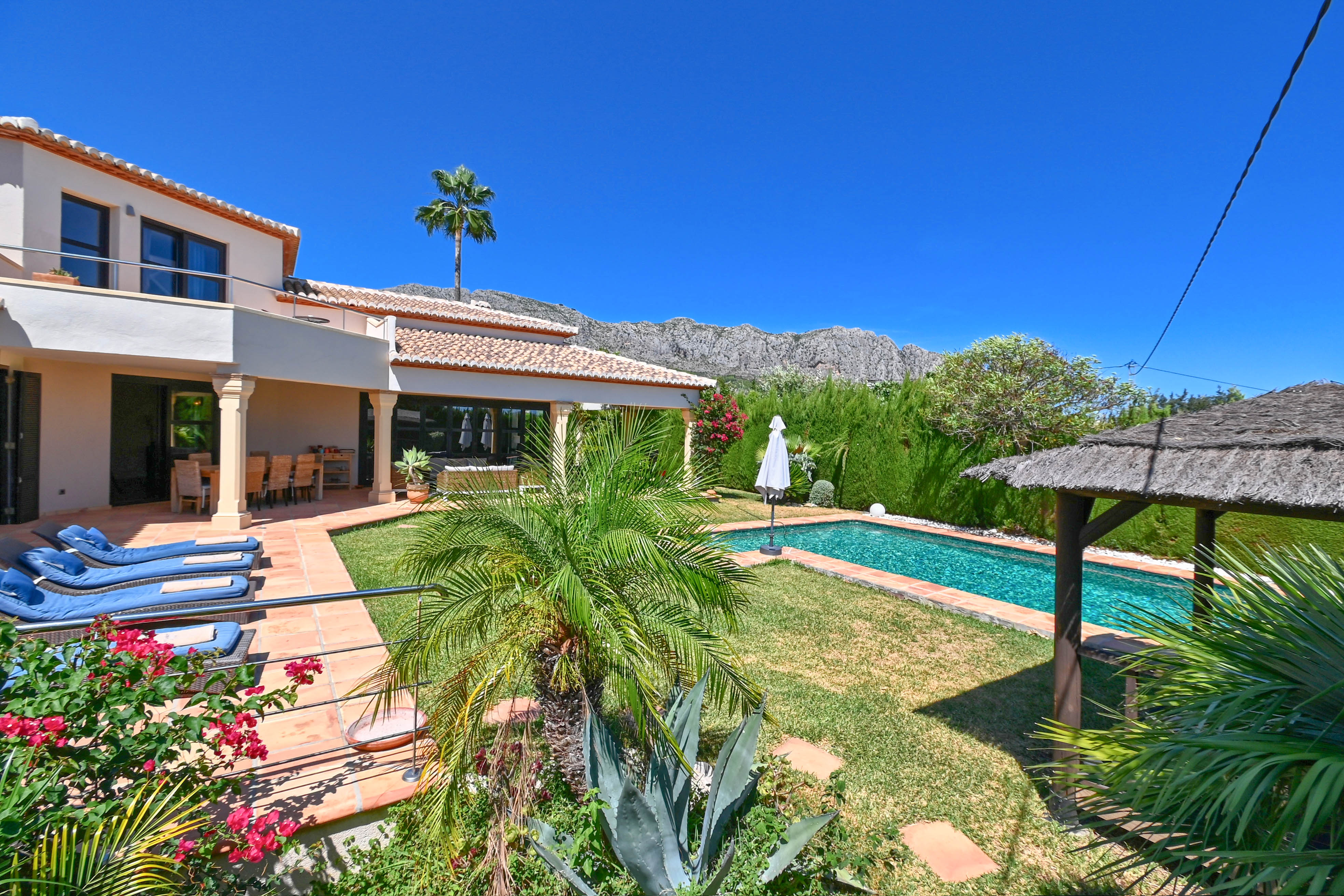 Vue sur le jardin avec piscine rectangulaire et gazebo en chaume, entouré d'une végétation luxuriante et de montagnes.