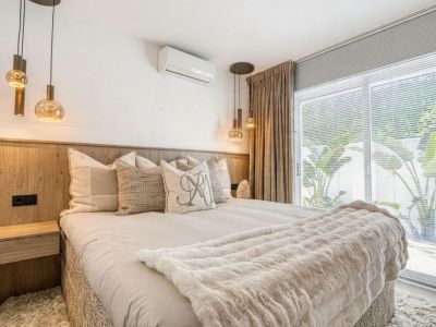 Chambre avec tête de lit en bois, suspensions en verre et rideaux beiges près d'une fenêtre.