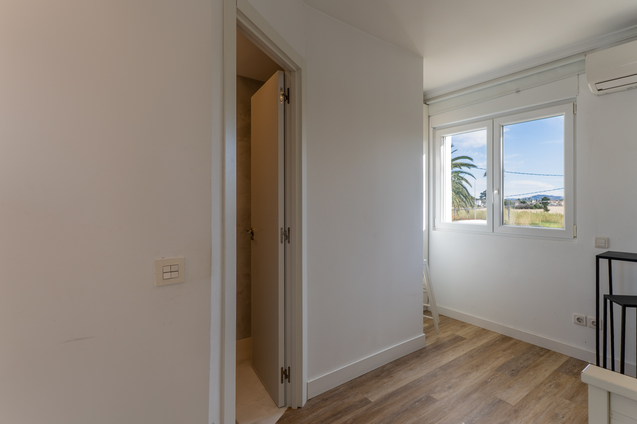 Chambre lumineuse avec plancher imitation bois et vue sur l'extérieur. Porte entrebâillée vers la salle de bain.