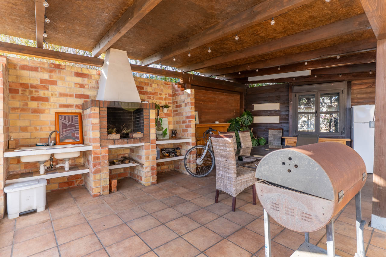 Espace barbecue extérieur avec four en brique, double évier et coin repas sous un toit en bois avec éclairage d'ambiance.