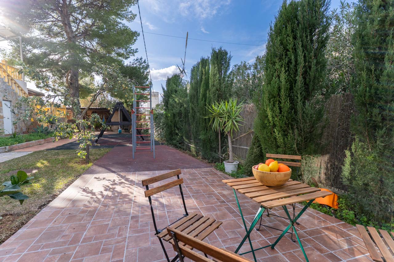 Terrasse avec table en bois et corbeille de fruits, entourée de verdure et d'une aire de jeux.