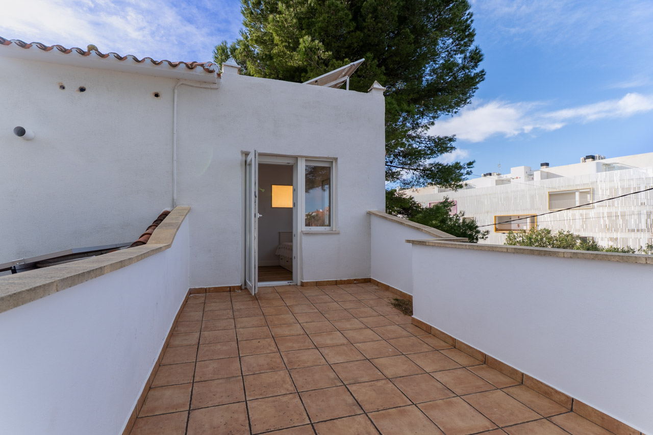 Terrasse carrelée avec murs blancs. Porte ouverte sur une chambre lumineuse.