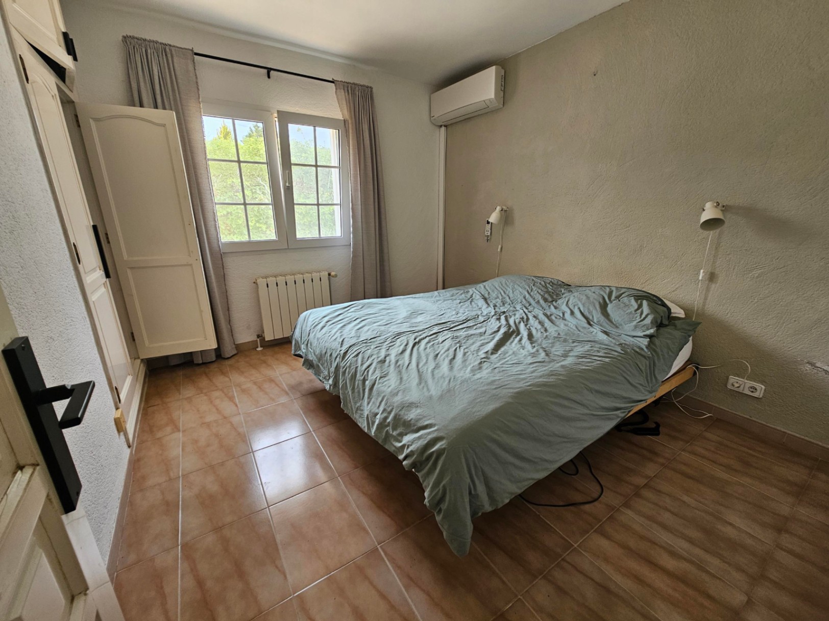 Chambre avec lit double, armoire blanche et sol en carrelage couleur terre cuite.