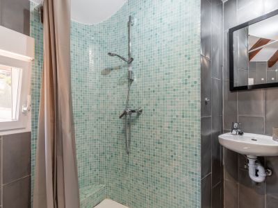 Détail de salle de bain avec douche en mosaïque turquoise, lavabo suspendu et murs carrelés gris.
