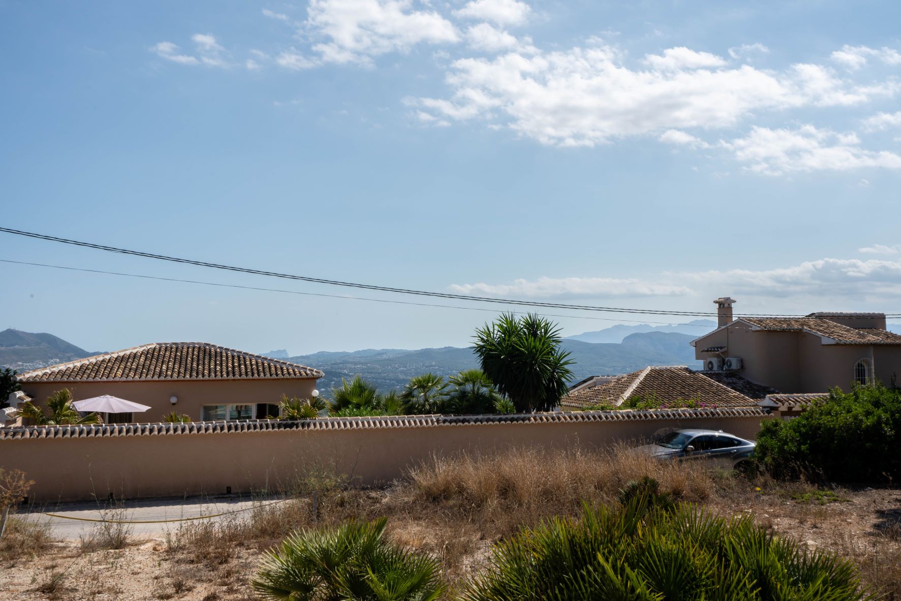Vue de la parcelle avec les maisons voisines, la végétation et les montagnes en arrière-plan sous un ciel partiellement nuageux.