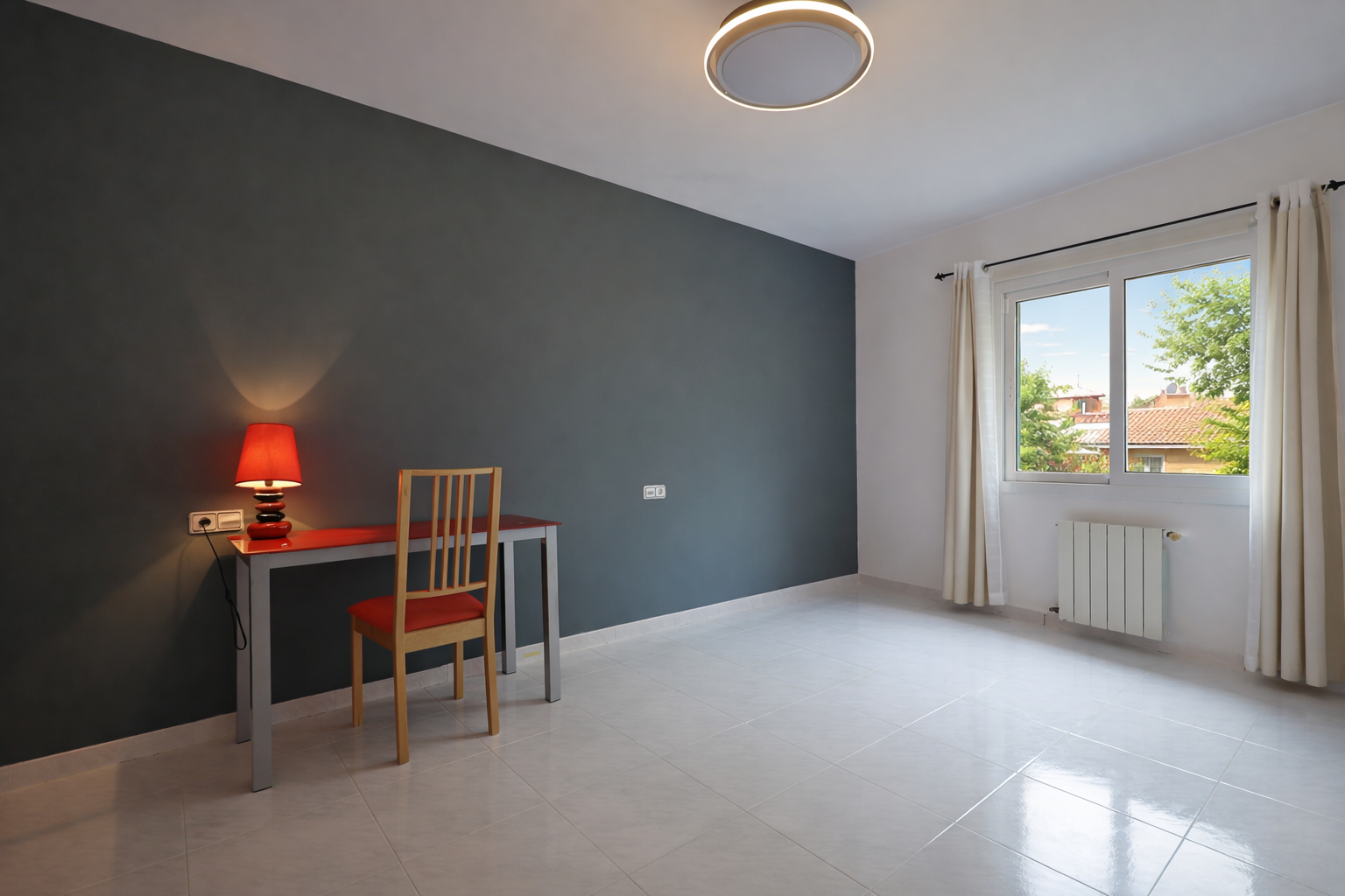 Chambre lumineuse avec bureau rouge et mur gris plomb, carrelage crème et plafonnier circulaire.