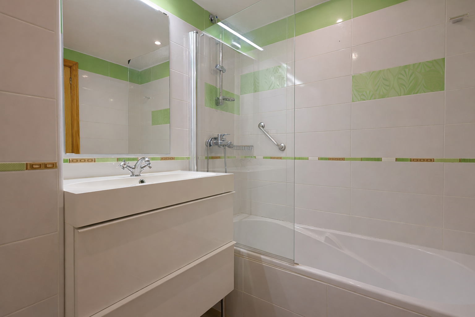 Salle de bain avec lavabo moderne, carrelage blanc à détails verts et douche avec paroi en verre.