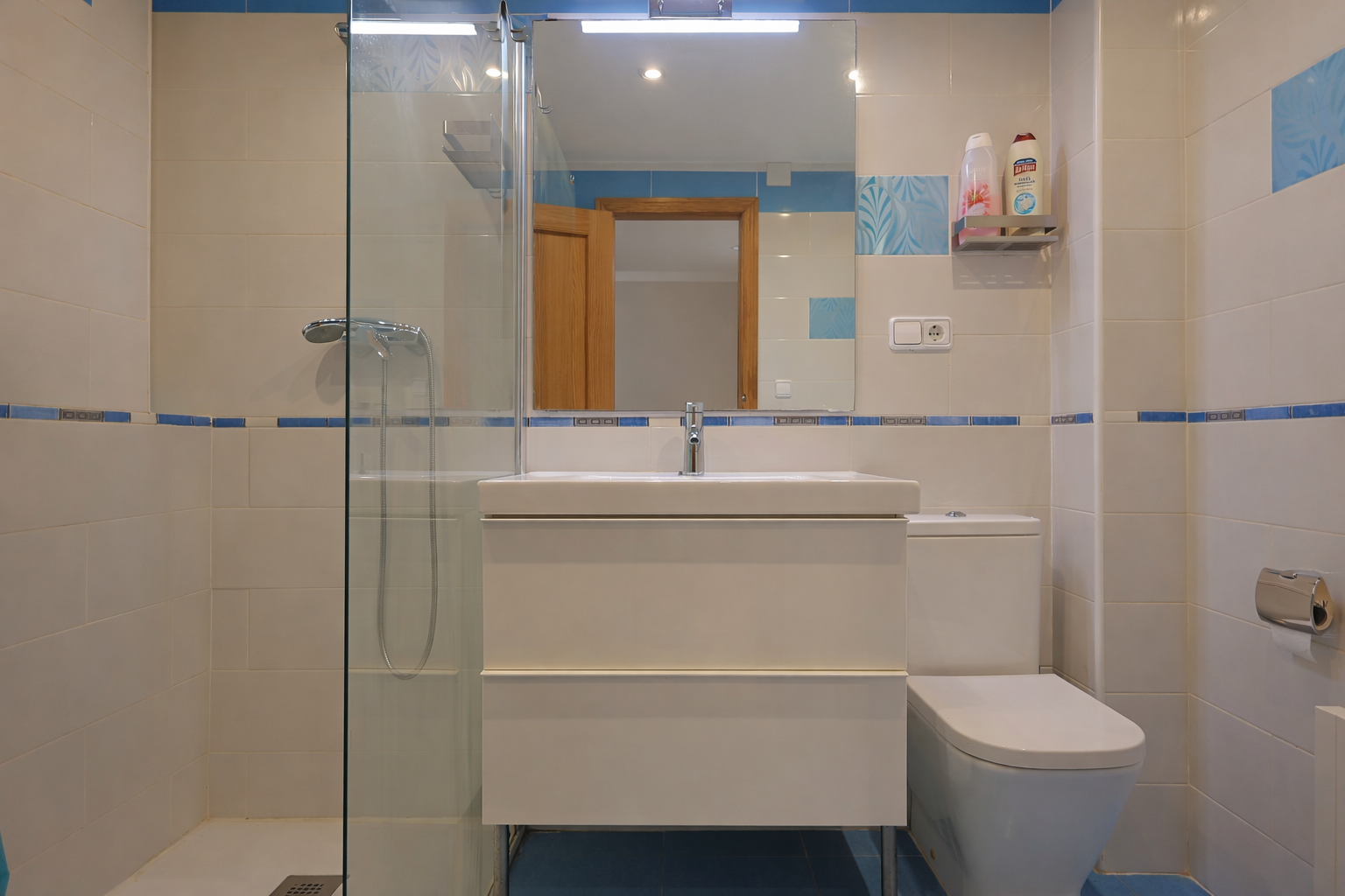 Salle de bain carrelée blanc avec détails bleus, miroir, meuble-lavabo moderne et douche à paroi de verre.