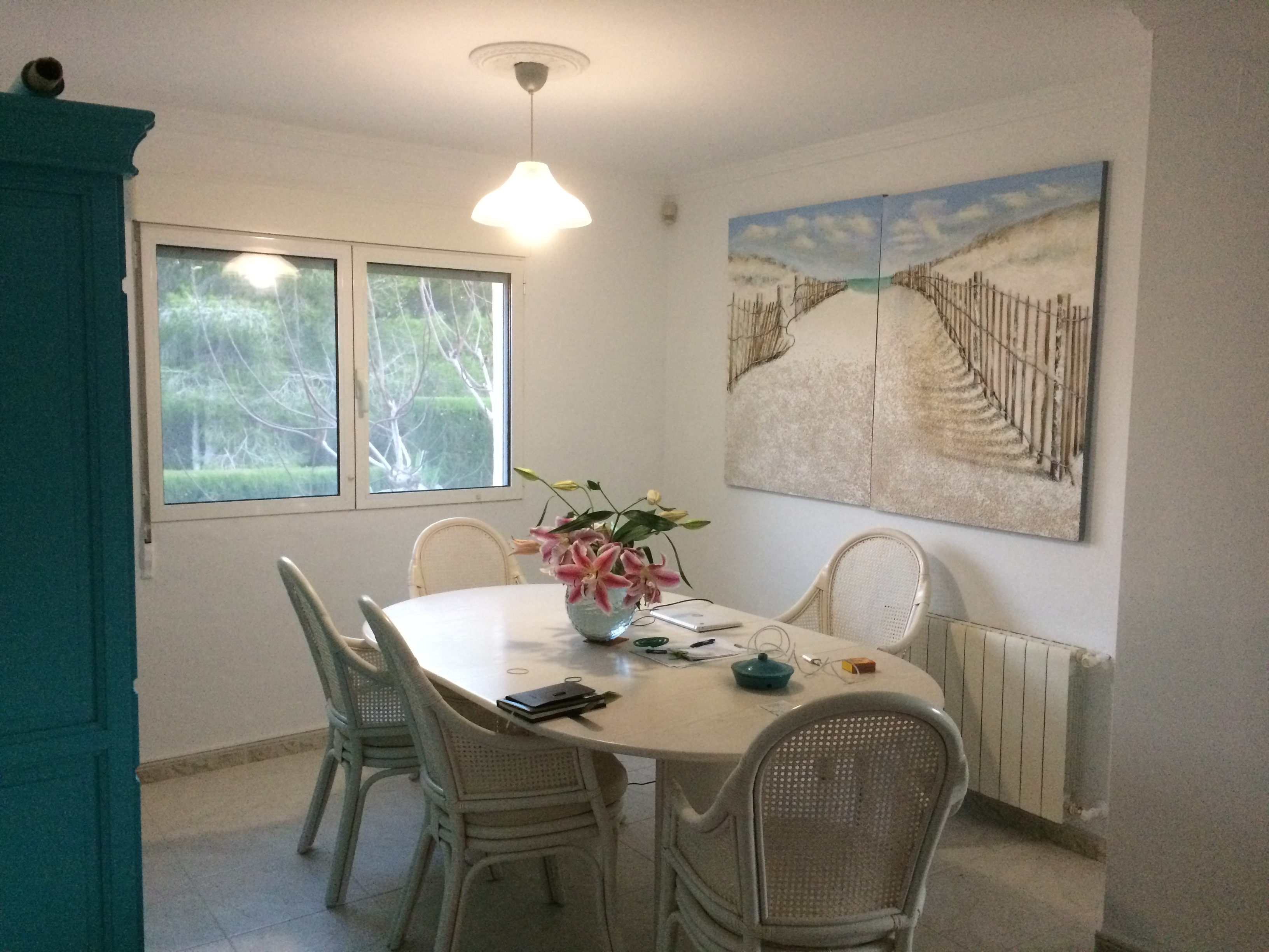 Salle à manger lumineuse avec table ovale en pierre, chaises en osier et tableau sur le thème de la plage.
