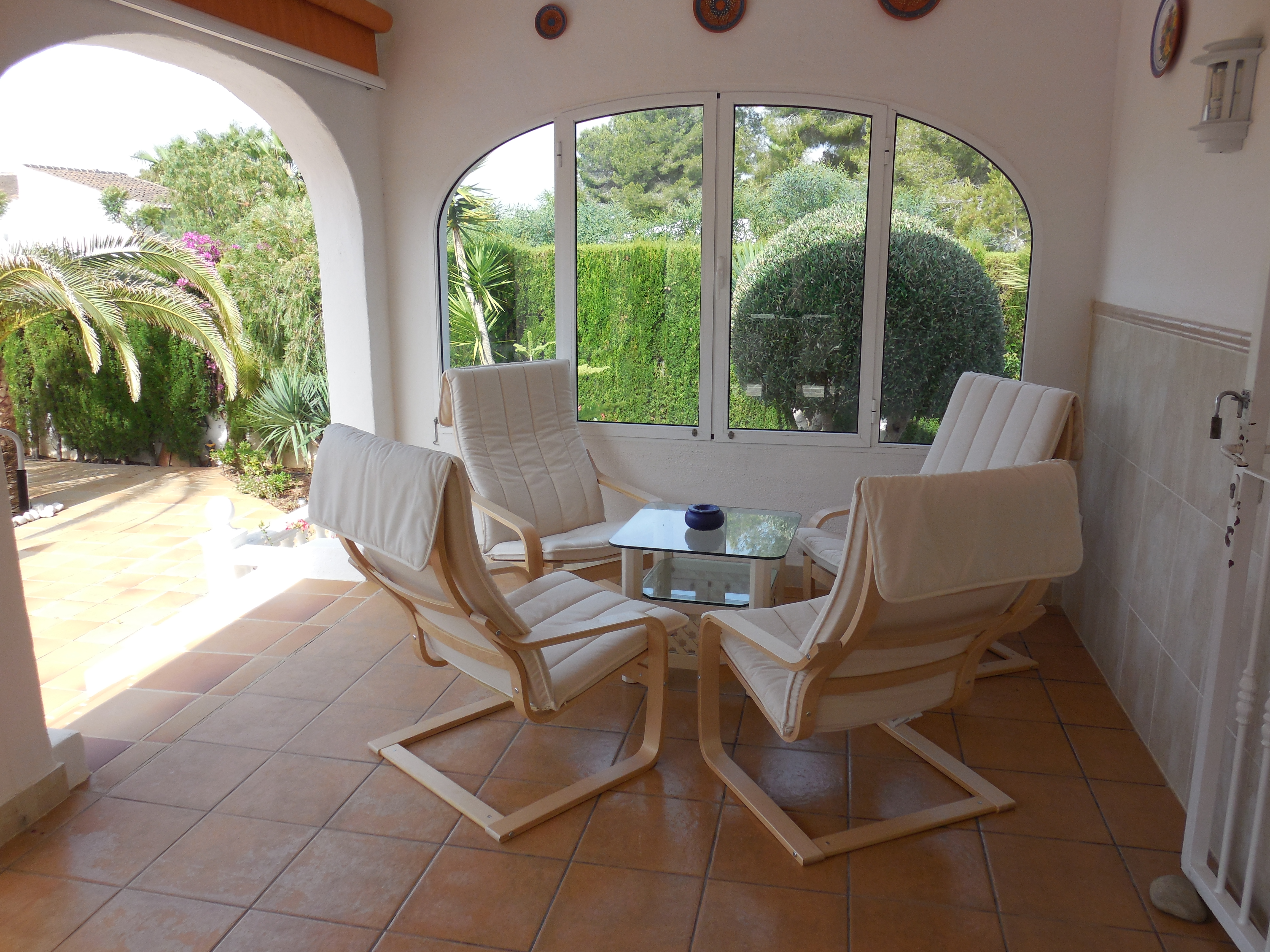 Terrasse couverte avec quatre fauteuils en bois clair et coussins blancs, table en verre et vue sur le jardin.