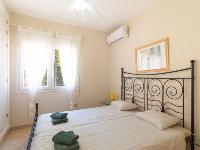 Chambre lumineuse avec lits jumeaux, tête de lit en fer forgé et vue sur l'extérieur à travers des rideaux blancs.