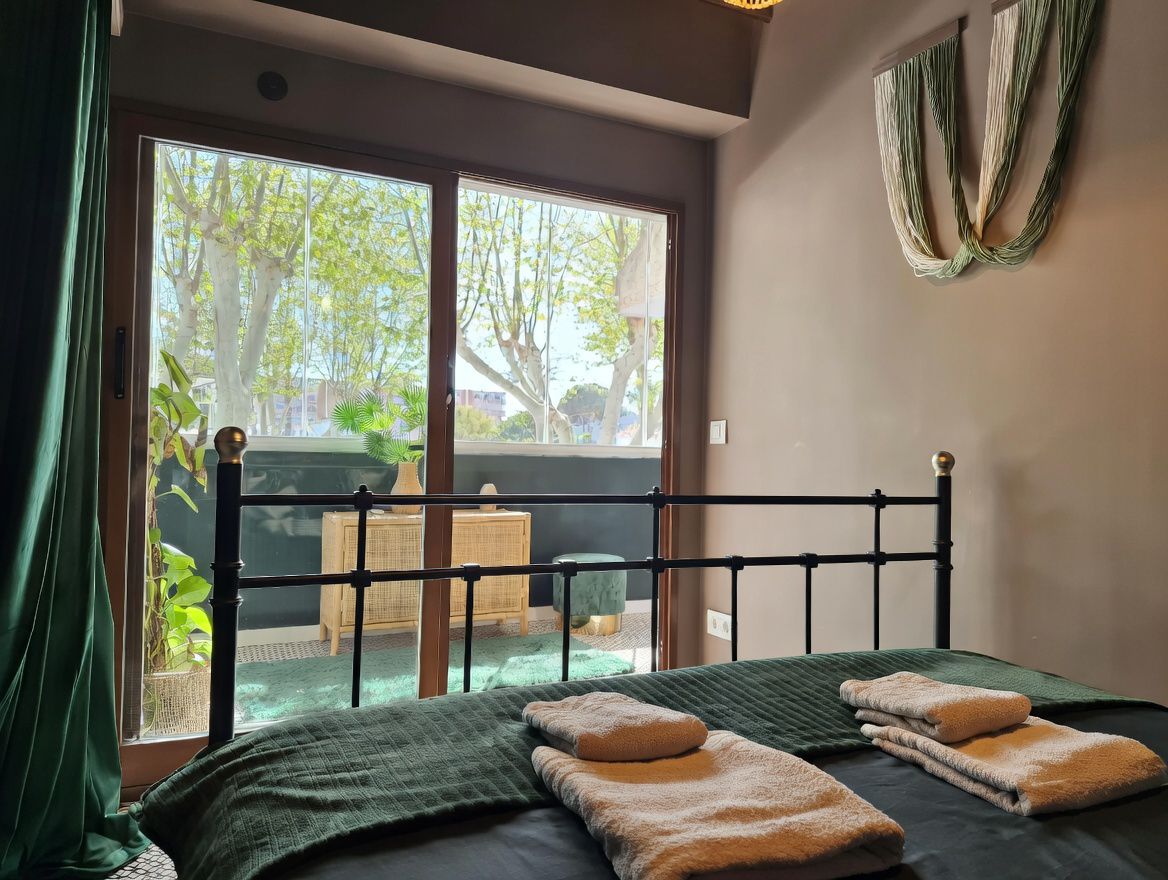 Chambre avec lit en fer forgé, balcon avec vue arborée et décor moderne dans des tons neutres et verts.