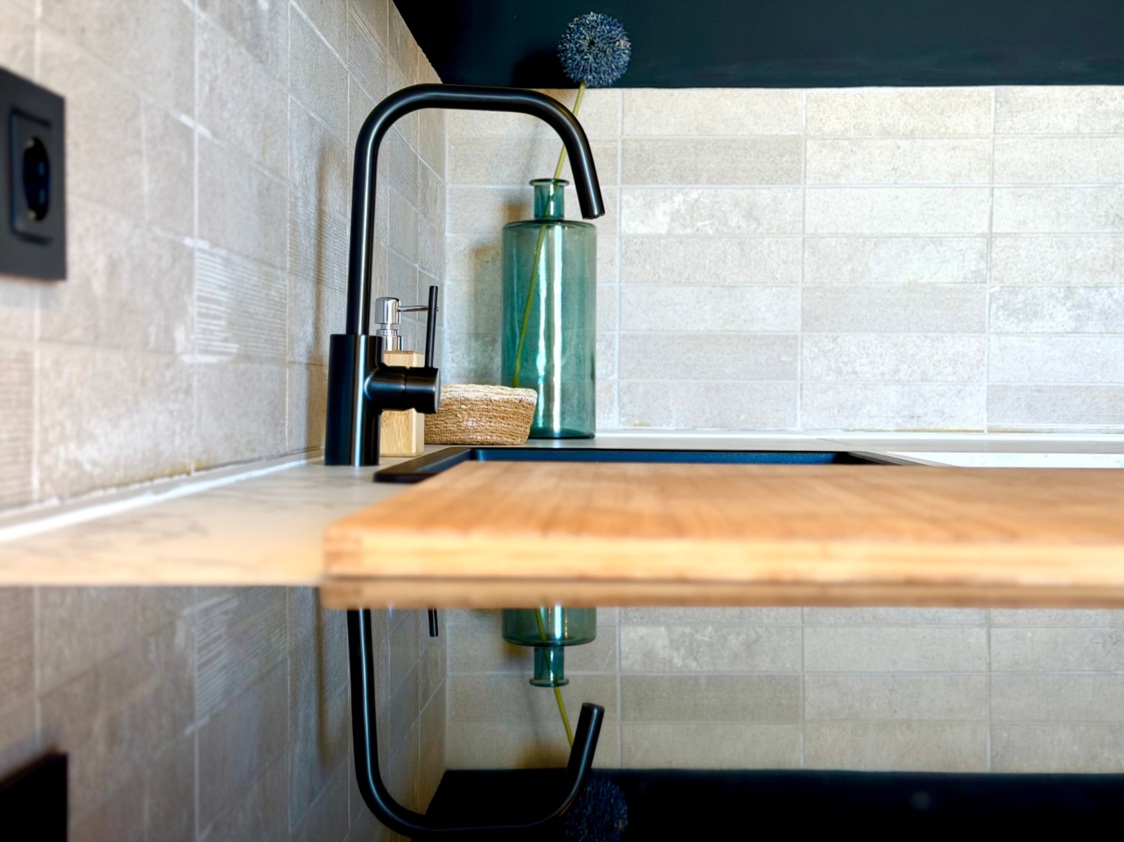Détail de la cuisine avec robinet noir, planche à découper en bois et carreaux couleur crème.