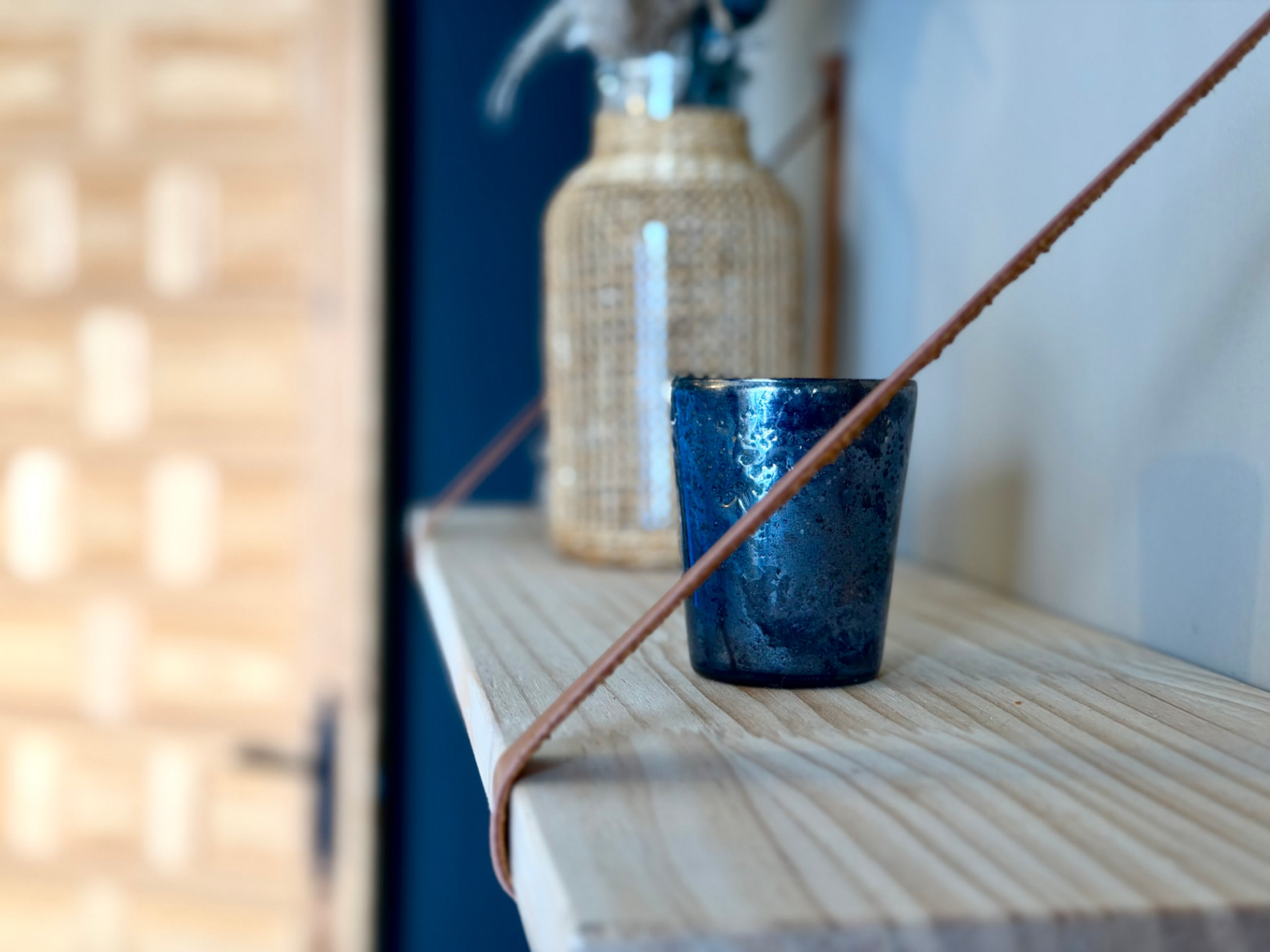 Gros plan d'une étagère en bois clair avec décor : un verre bleu texturé et une bouteille en verre avec un manchon en osier.