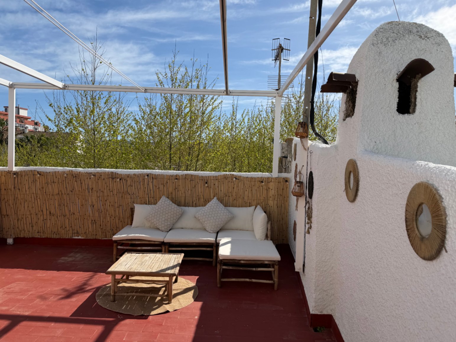 Terrasse avec sol rouge, mur blanc décoré, canapé en bambou et vue sur la verdure.