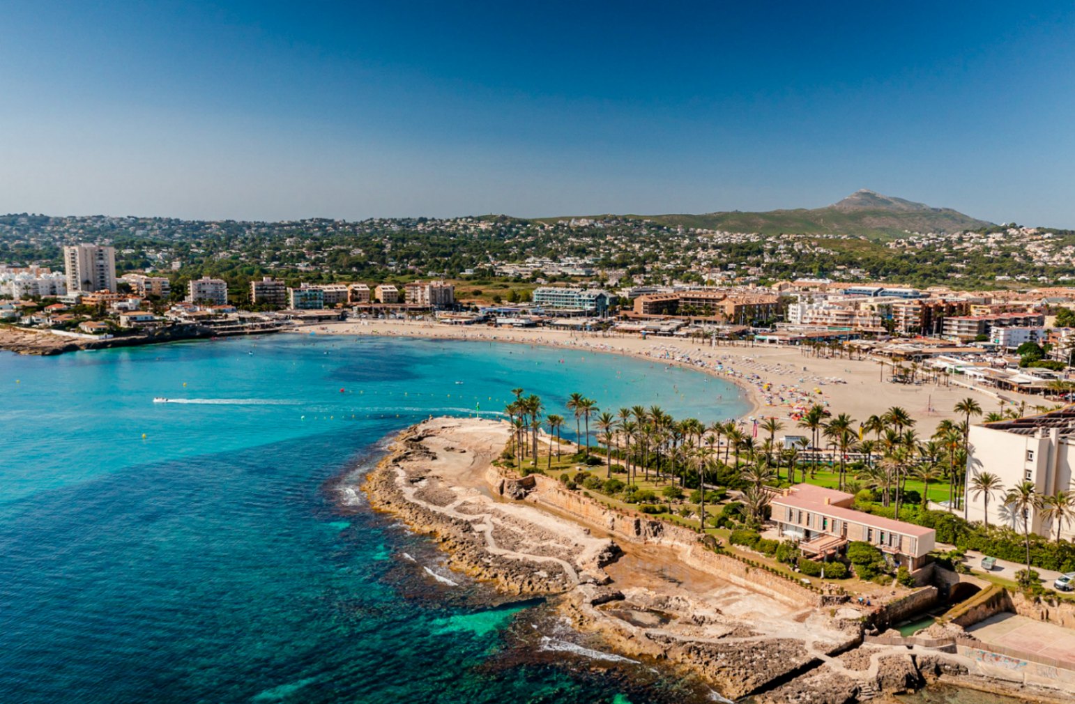 Vue aérienne de la Playa del Arenal à Jávea, avec des eaux turquoise et des bâtiments côtiers.