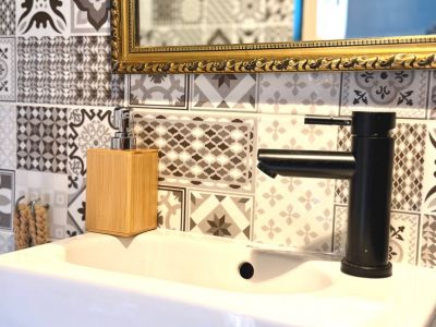 Gros plan d'un lavabo avec robinet noir mat et distributeur de savon en bambou. Carrelage décoratif au mur et miroir à cadre doré.