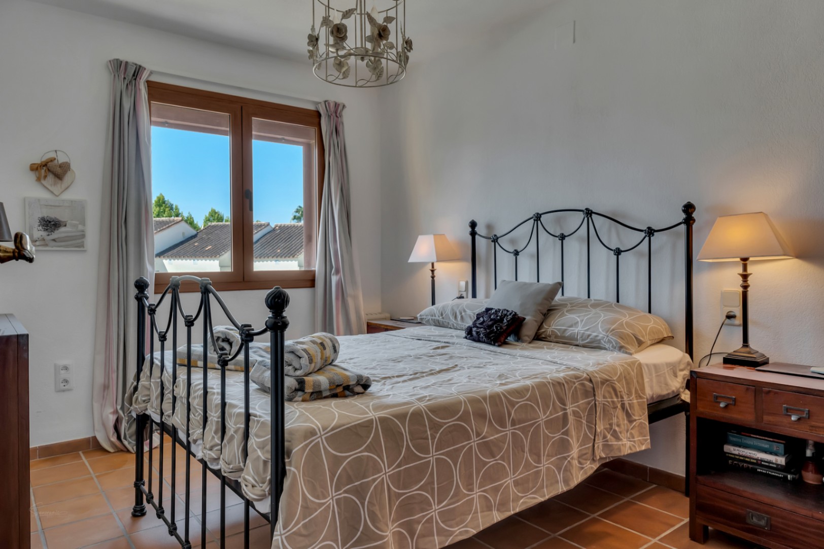 Chambre lumineuse avec lit en fer forgé, fenêtre avec vue extérieure et sol en terre cuite.