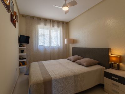 Chambre à coucher avec lit double, tête de lit rembourrée et table de chevet avec lampe. La fenêtre est habillée de voilages, complétée par un ventilateur de plafond et une télévision murale.