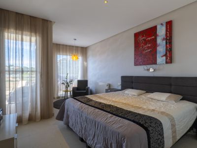 Chambre lumineuse avec lit double, tête de lit grise capitonnée et accès direct à une terrasse. Le sol carrelé clair et un luminaire suspendu décoratif complètent l'éclairage naturel.