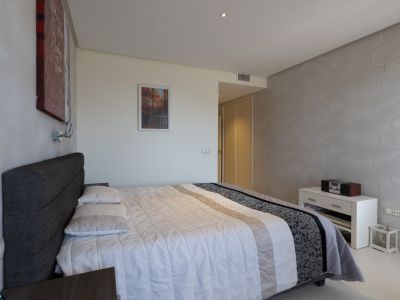 Chambre principale avec un lit double, une tête de lit capitonnée grise et une couette blanche. Un mur décoratif et des armoires encastrées sont visibles.