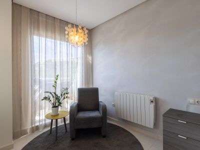 Coin lumineux avec fauteuil gris, table d'appoint jaune et plante. Grande fenêtre voilée, luminaire suspendu décoratif et radiateur mural électrique.