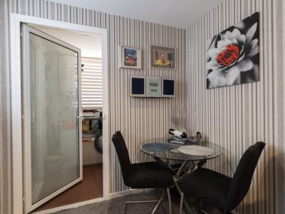 Coin repas intime avec table en verre et chaises noires, murs recouverts de papier peint rayé et une œuvre florale. Une porte ouverte mène à une buanderie éclairée.