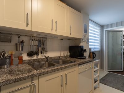 Cuisine fonctionnelle avec armoires crème, plan de travail en granit moucheté avec double évier en inox et lave-vaisselle blanc intégré. Lumière naturelle par la fenêtre à stores.