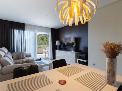 Espace de vie et salle à manger ouverts avec un lustre en spirale distinctif, un canapé sectionnel gris clair et une table blanche. Une porte vitrée mène à un balcon verdoyant.