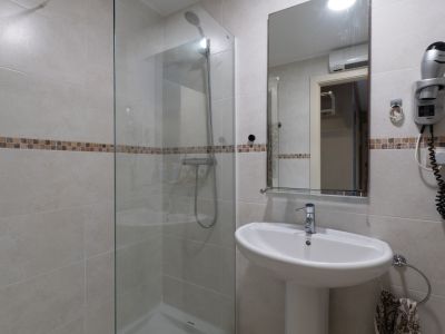 Salle de bain avec douche vitrée, carrelage clair et bande décorative en mosaïque. Lavabo sur pied, miroir et sèche-cheveux mural inclus.