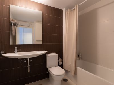 Salle de bain fonctionnelle avec lavabo mural, toilettes et baignoire avec rideau de douche. Elle présente des carreaux marron foncé contrastant avec des carreaux muraux clairs.