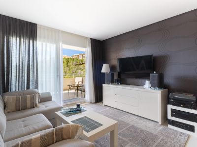 Salon spacieux avec canapé sectionnel gris clair, table basse blanche et meuble TV devant un mur texturé. Portes coulissantes vers un balcon avec vue sur la colline.