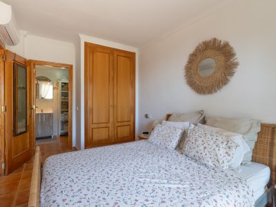 Chambre à coucher avec lit à tête de lit en osier, armoire encastrée en bois clair et sol en terre cuite. Un miroir en fibres naturelles décore le mur, avec vue sur une salle de bain.