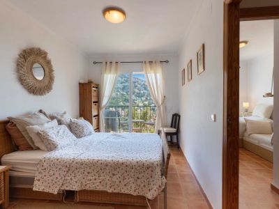 Chambre avec lit en osier, carrelage en terre cuite et accès direct à un balcon offrant une vue sur la colline. Miroir en raphia et meuble en bois.