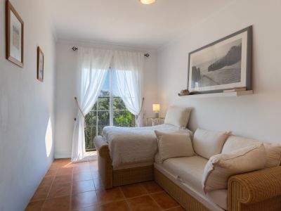 Chambre d'amis lumineuse avec sol en terre cuite, lit simple et canapé en rotin. La fenêtre avec voilages offre une vue sur la verdure environnante.