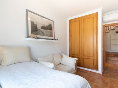 Chambre secondaire lumineuse avec lit simple, canapé d'appoint, armoire encastrée en bois et étagère murale. Carrelage en terre cuite et accès à un couloir avec escalier.