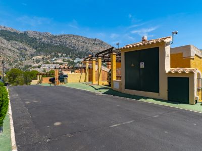 Chemin d'accès résidentiel avec bâtiment de services jaune, caméra de sécurité et pergola, devant une chaîne de montagnes verdoyante parsemée d'habitations sous un ciel bleu clair.