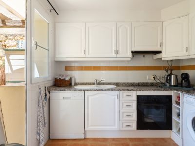 Cuisine fonctionnelle avec armoires blanches, plan de travail effet granit et appareils intégrés. Une fenêtre ouverte donne sur un patio extérieur avec murs en pierre et pergola en bois.