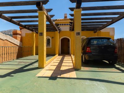 Entrée extérieure avec façade en stuc jaune et pergola en bois couvrant une zone de stationnement verte. Comprend une porte cintrée, un chemin carrelé et une voiture noire garée à l'ombre.
