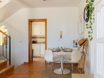 Espace repas lumineux avec table ronde et chaises modernes, sol en terre cuite et cuisine compacte adjacente. Un escalier en bois avec fenêtre cintrée est visible.