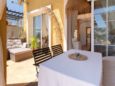 Grande terrasse extérieure avec sol carrelé, canapé de salon en rotin et table à manger, offrant des vues partielles sur les collines.