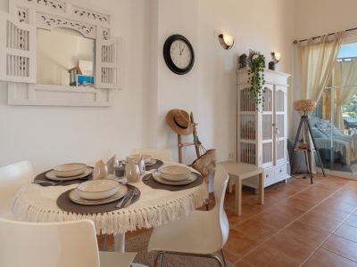 Salle à manger lumineuse avec table ronde dressée, chaises blanches et miroir mural décoratif en bois. Le sol en terre cuite mène à une terrasse avec vue sur les collines.