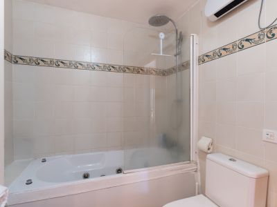 Salle de bain complète avec baignoire d'angle à jets et douche. Murs carrelés beiges avec frise décorative et toilettes. Étagère de rangement visible sur le côté.